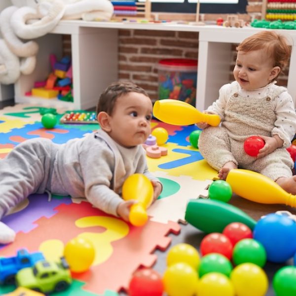 Two adorable babies playing with balls and bowling pin sitting on floor at kindergarten